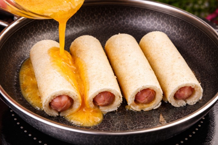 pouring egg mixture over bread roll sausages in a frying pan